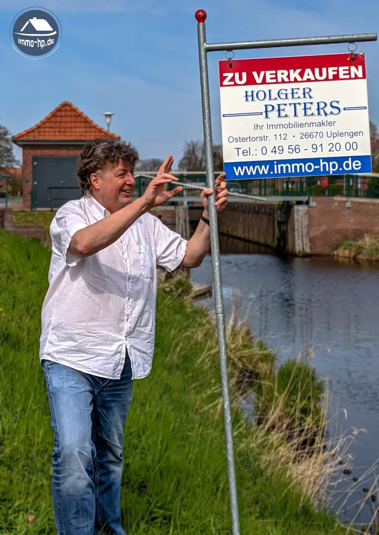 Holger Peters, Immobilienmakler für Augustfehn und Ostfriesland, mit Verkaufsschild an einer Fehn-Lage – persönliche Betreuung und regionale Marktkenntnis.